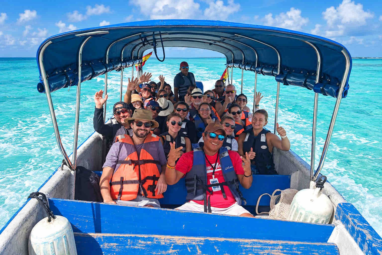Smiling travelers in life jackets waving aboard a covered boat in turquoise water.