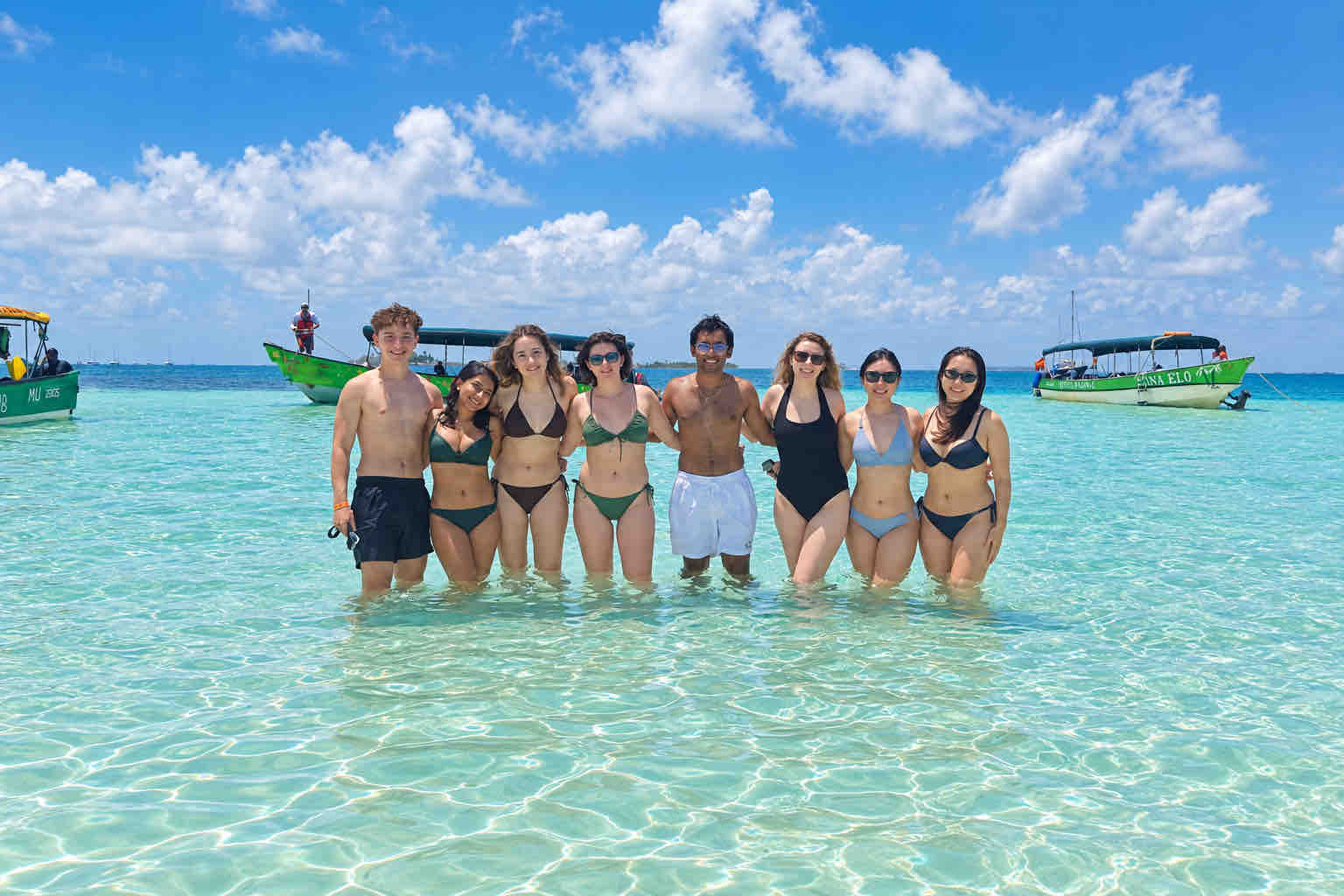 Eight travelers posing together in clear shallow water with boats behind them.