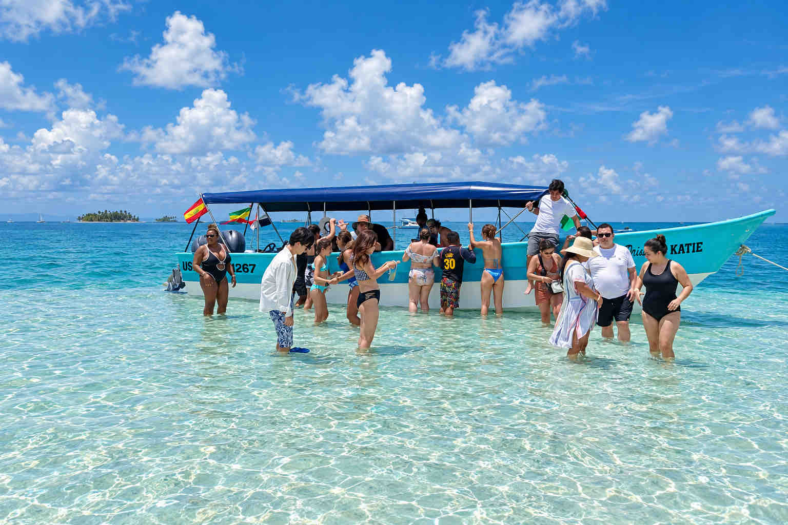 Travelers wading beside a turquoise tour boat in shallow Caribbean water.
