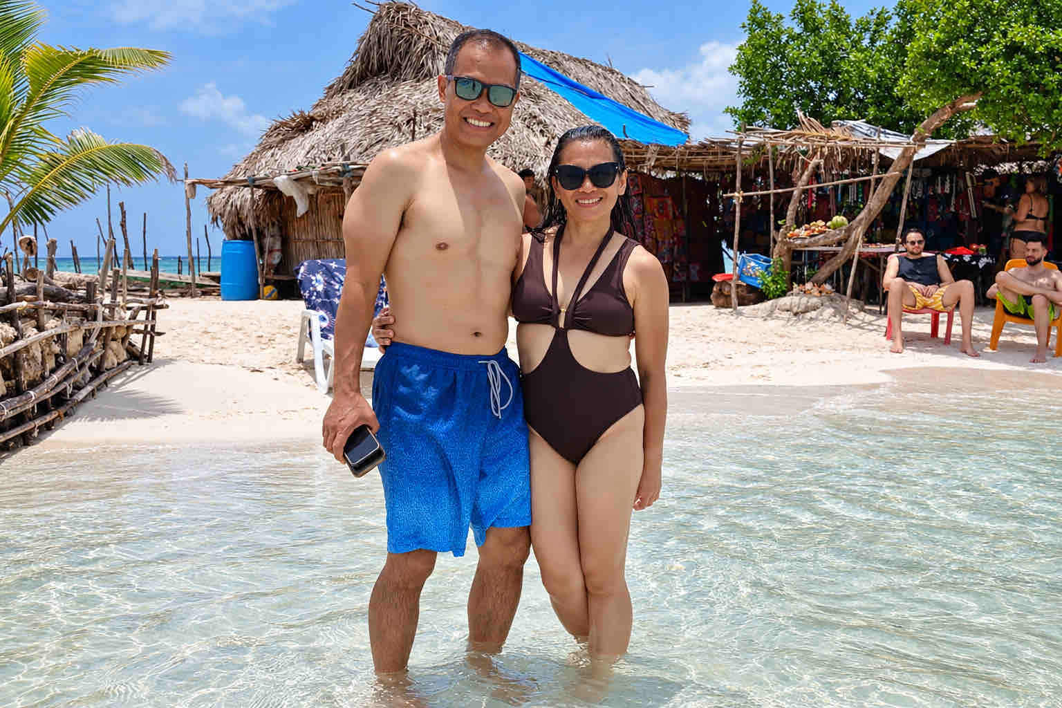 Two travelers standing in shallow water beside thatched island huts during a San Blas beach trip