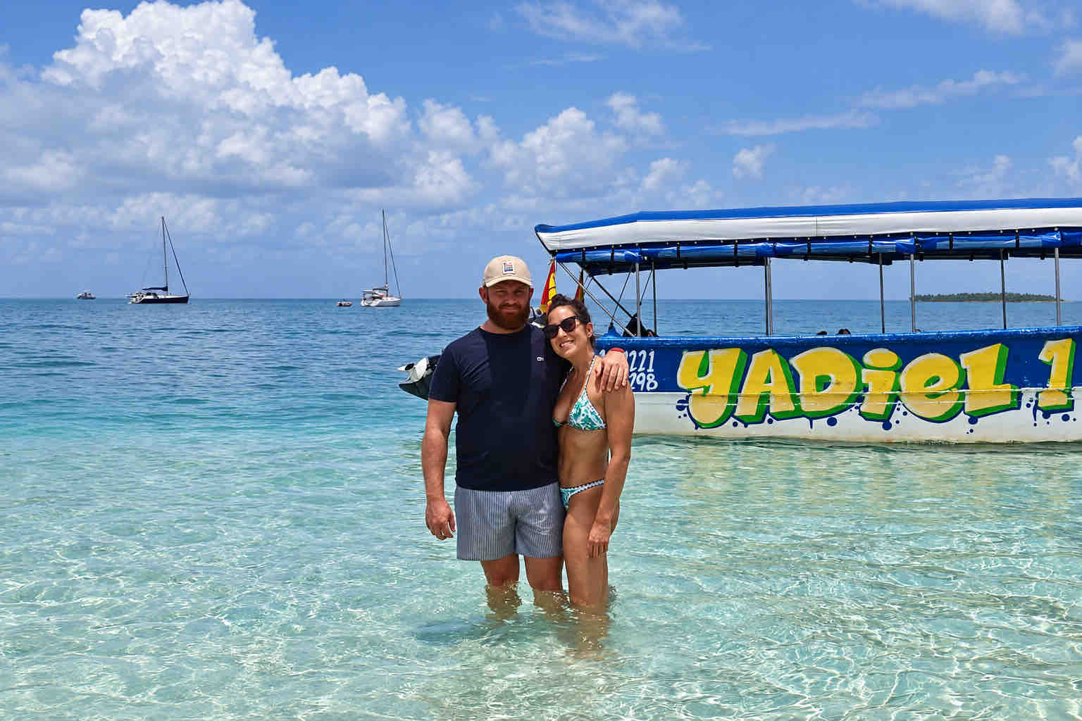 Two travelers standing in shallow water beside a covered tour boat during a San Blas beach trip