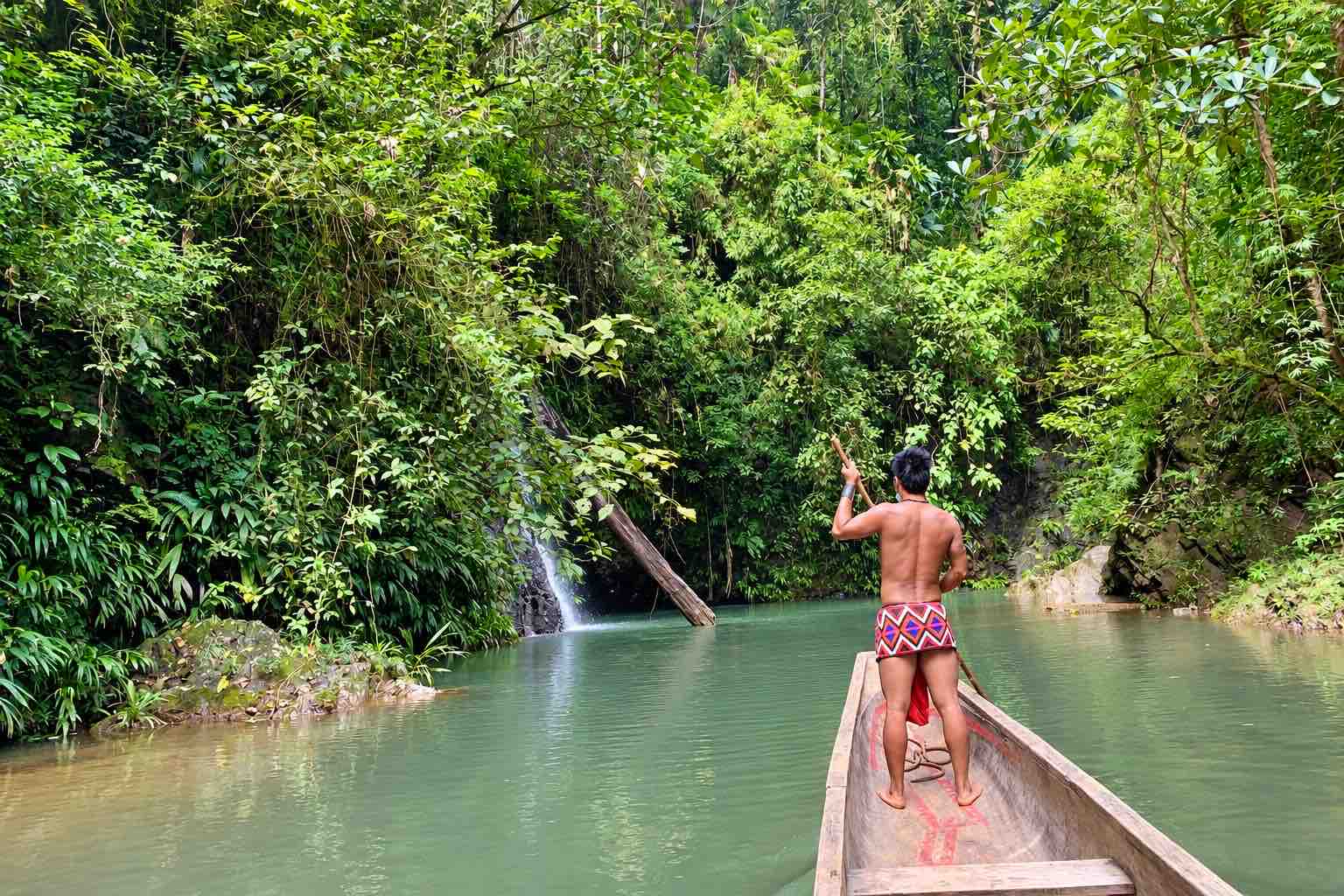 Emberá guide steering a wooden canoe through a narrow forest stream beside a small cascade