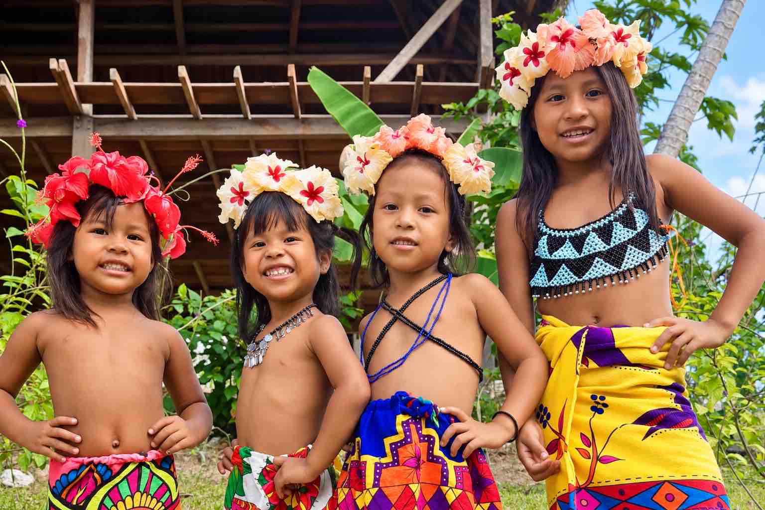Four Emberá children wearing flower crowns and colorful traditional clothing outside a village house