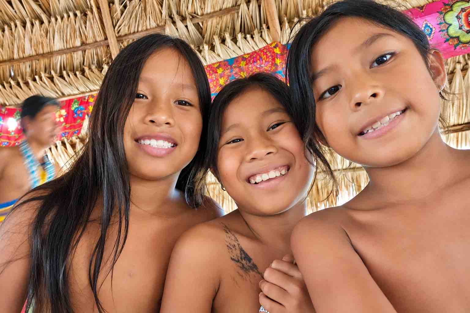 Three smiling Emberá children beneath a thatched roof with colorful textiles behind