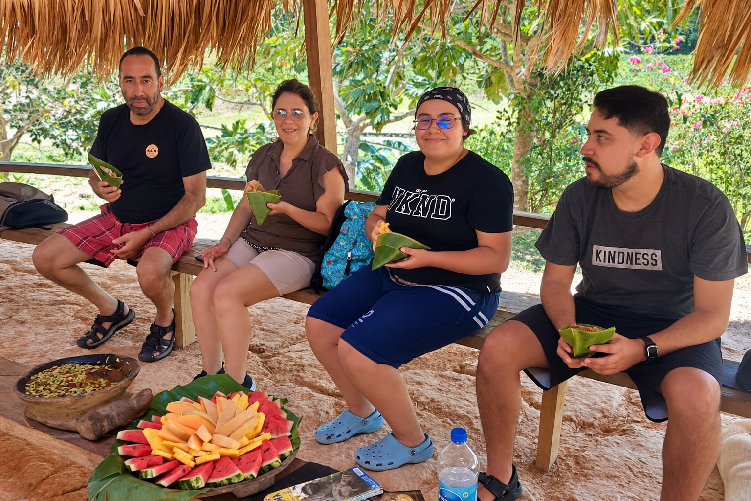 Four visitors eating leaf-wrapped meals beneath a thatched roof with a fresh fruit platter in front