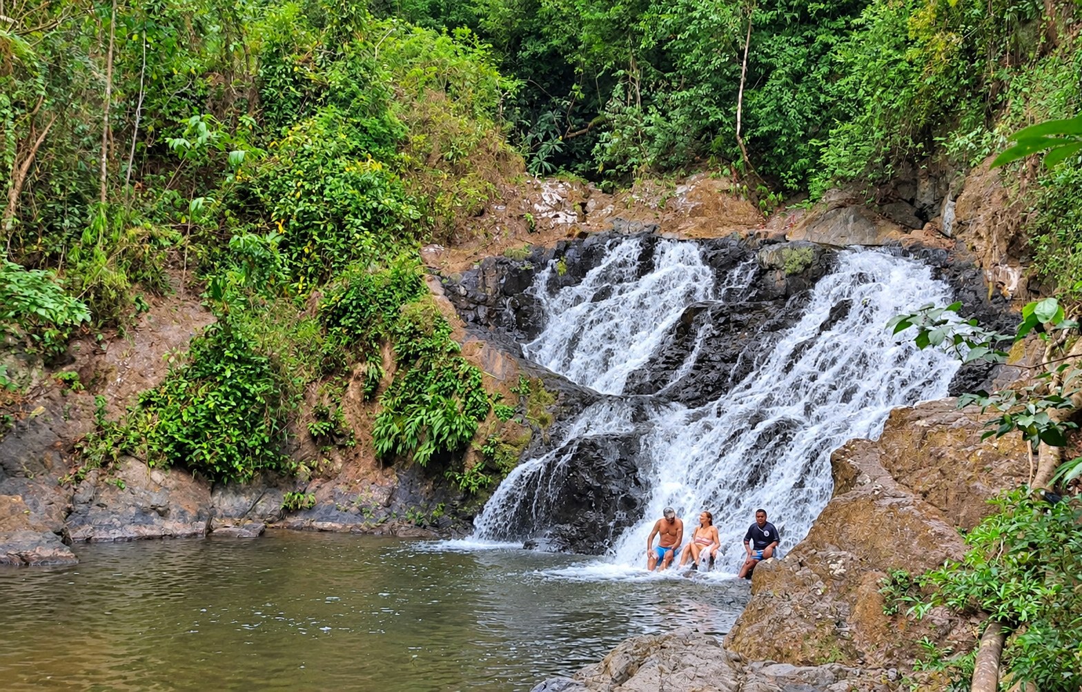 Three visitors sitting beneath a cascading waterfall surrounded by rainforest and rocks