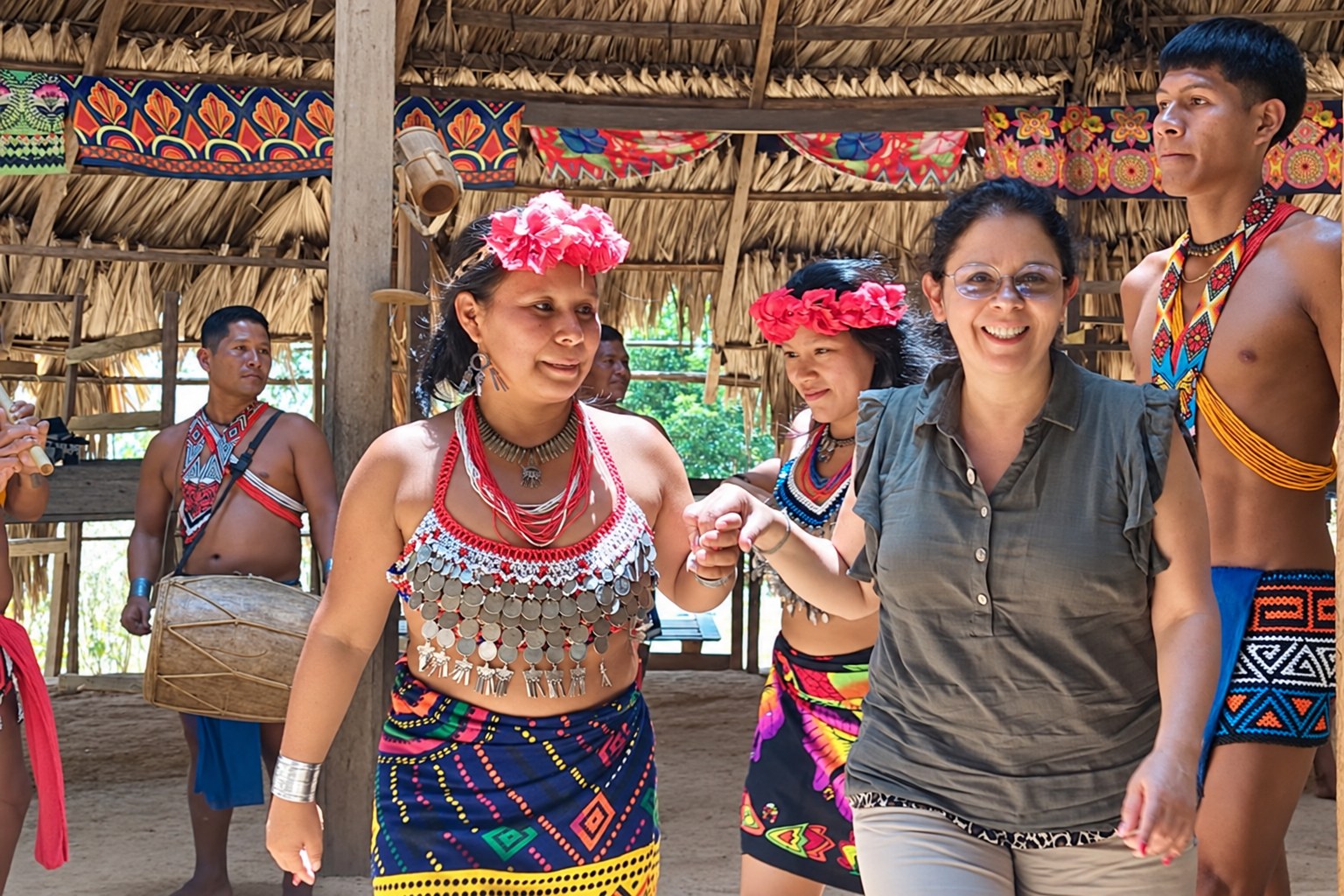 A visitor dancing with Emberá hosts in traditional clothing while a drummer plays beneath a thatched roof