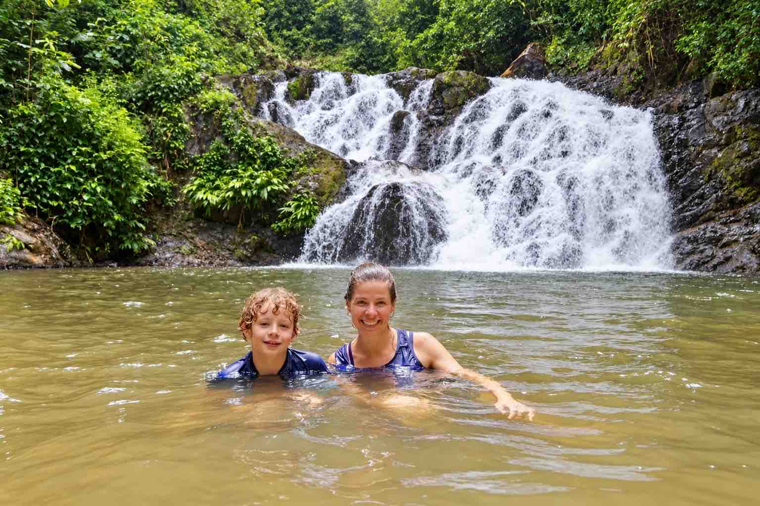 An adult and child swimming in a natural pool with a rainforest waterfall behind