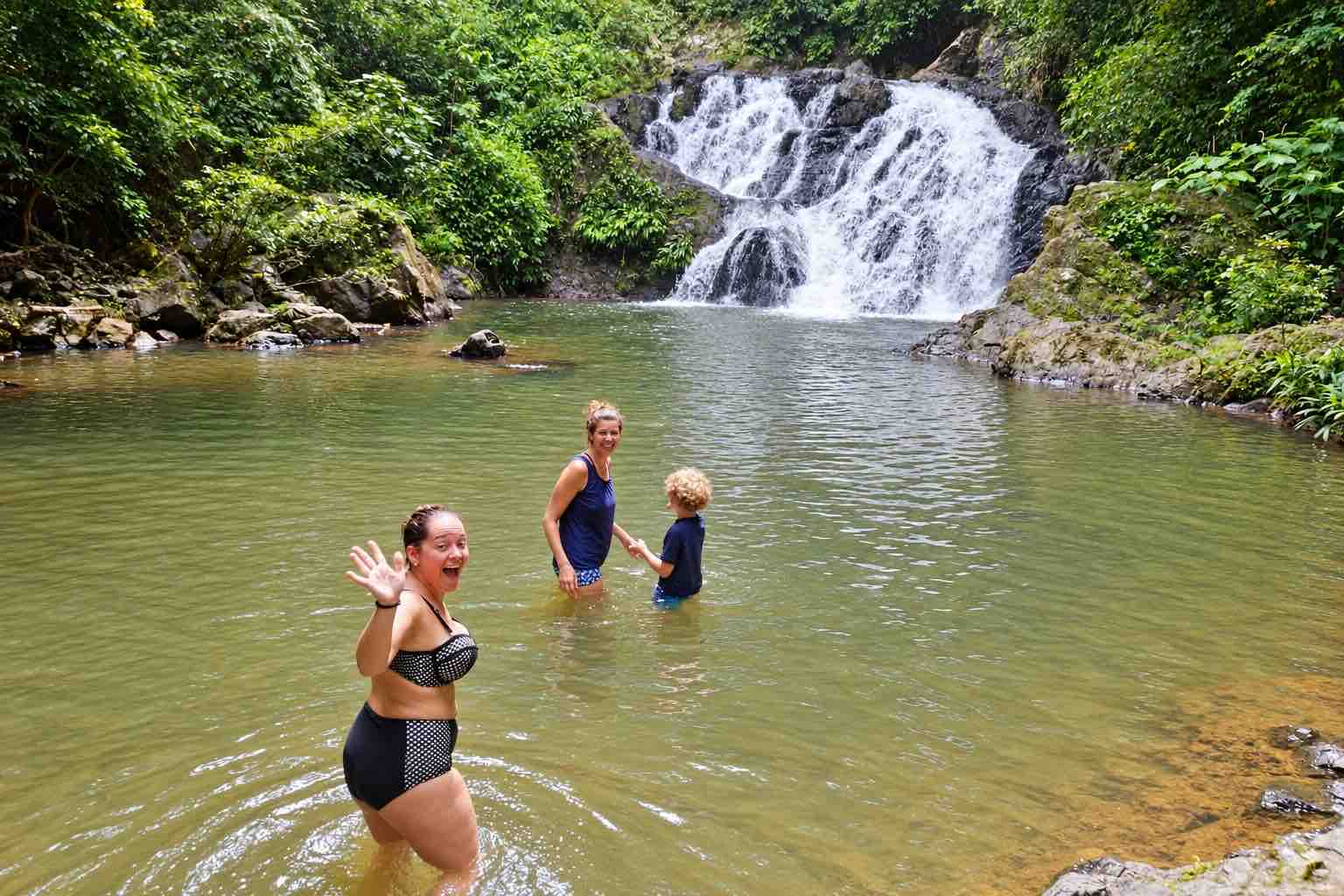 Three visitors standing in a natural pool surrounded by rainforest beneath a waterfall