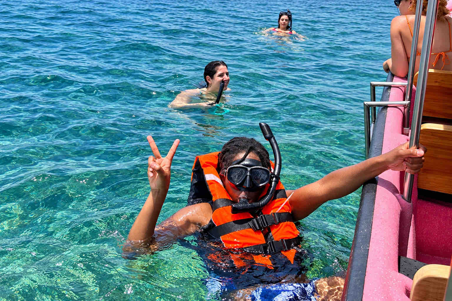Traveler wearing an orange life jacket and snorkel mask holding a boat ladder and making a peace sign.