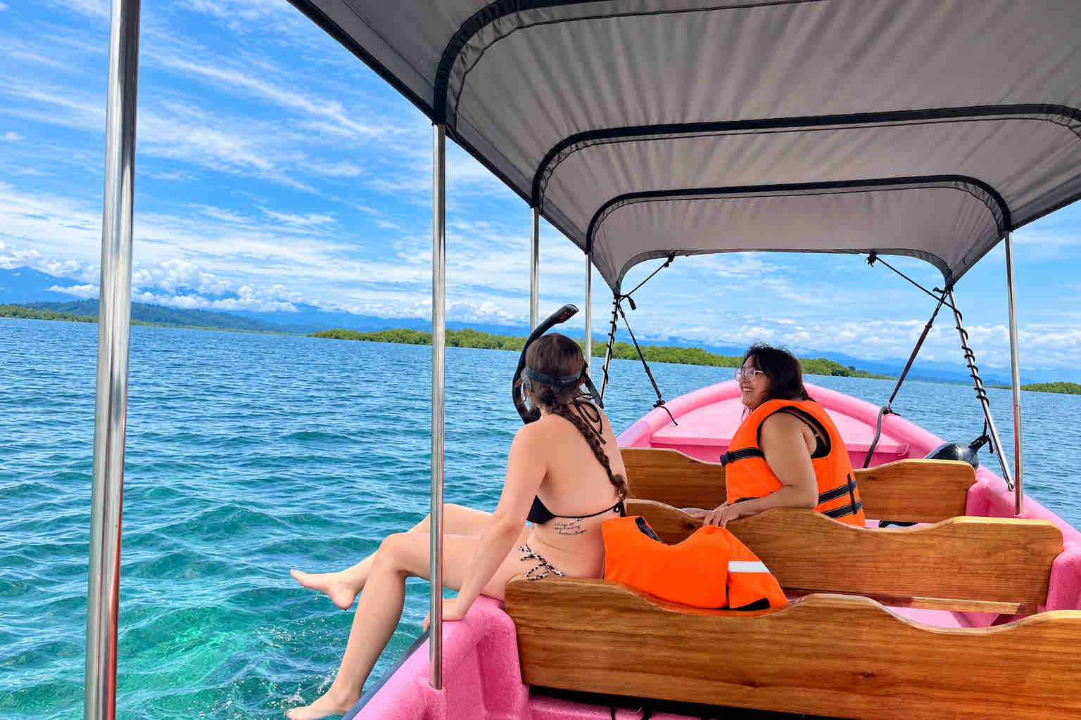 Traveler wearing a snorkel mask seated at the edge of a pink boat beside another passenger.