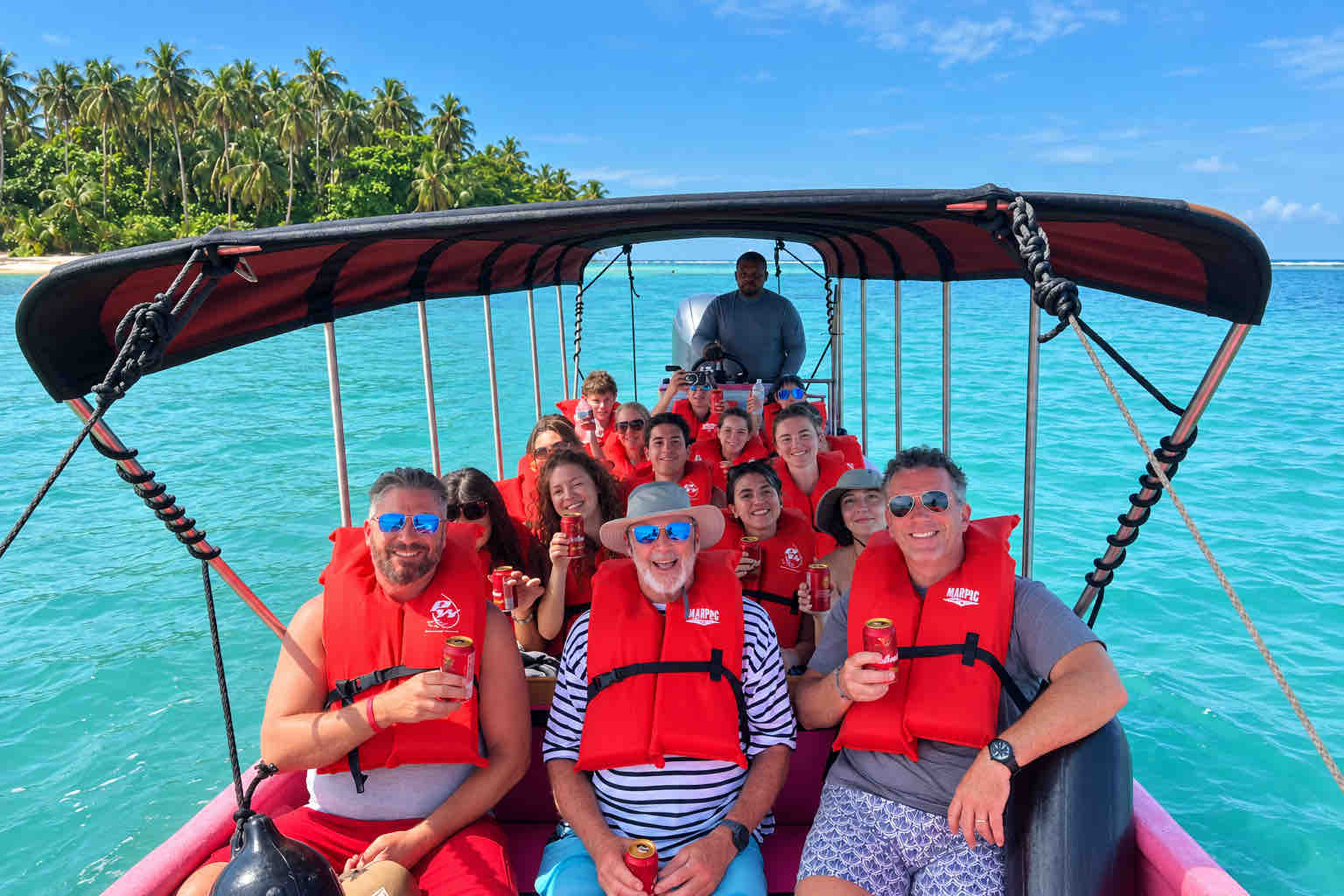 Smiling travelers wearing orange life jackets aboard a pink boat near a palm-lined island.