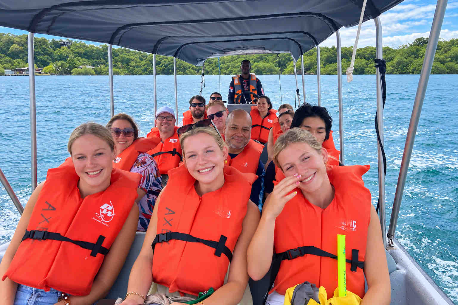 Smiling travelers wearing orange life jackets aboard a covered tour boat.