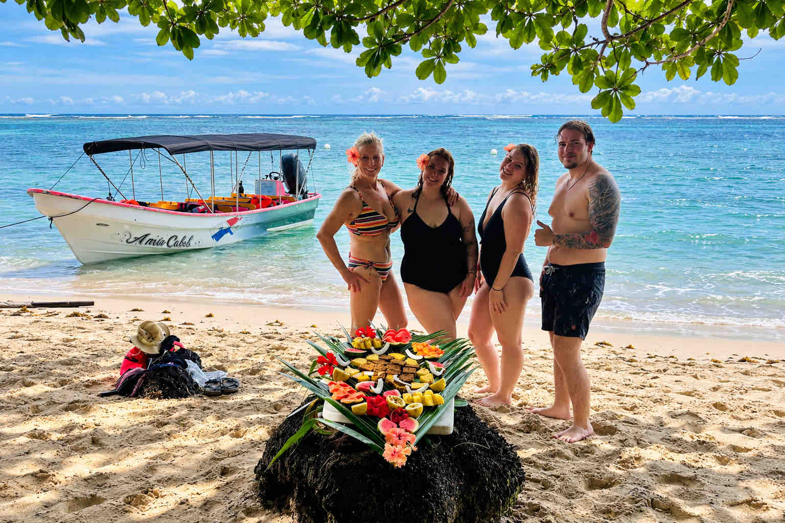 Four travelers beside a tropical fruit display on a beach, with a covered boat behind them.