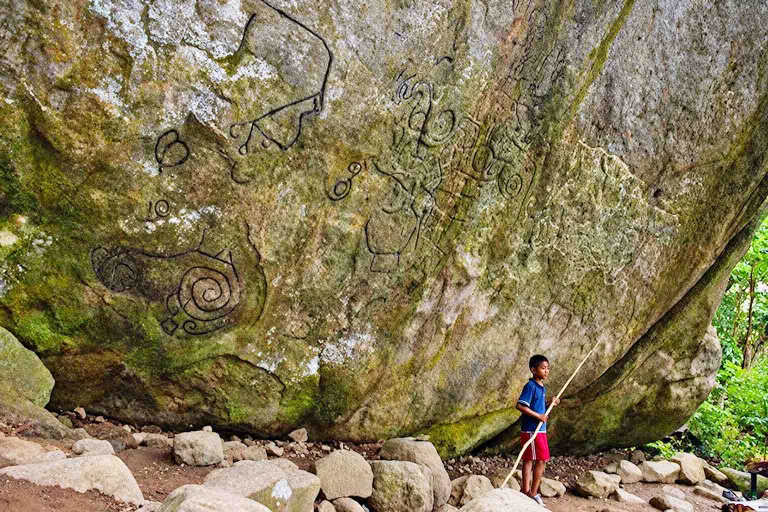 Person standing beside the large Piedra Pintada rock covered in carvings