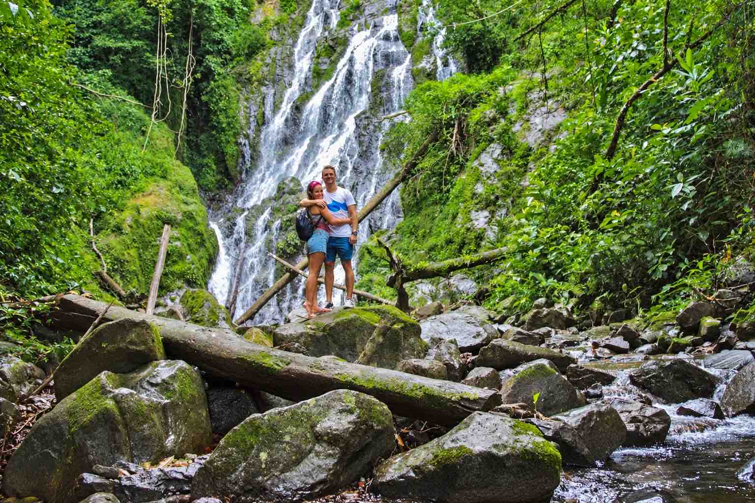 Two visitors standing on rocks in front of a forest waterfall