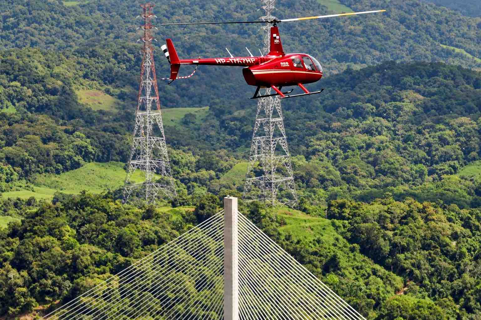 Panama helicopter tour: Red helicopter flying above forested hills and a cable-stayed bridge.