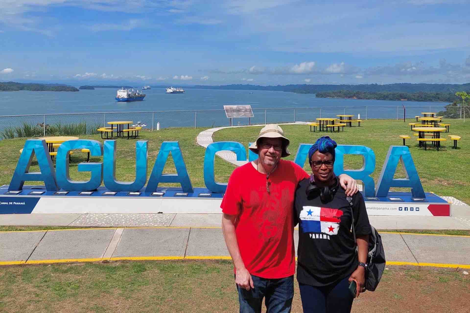 Couple posing beside the Agua Clara sign with canal waters behind them on the Ocean to Ocean Panama Canal Tour