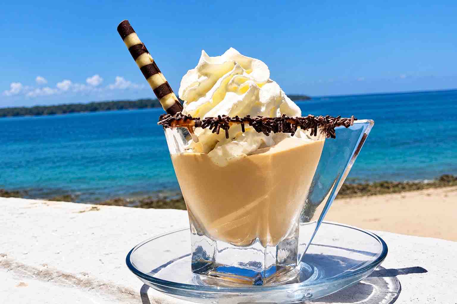 Mar y Oro Contadora – Cream-topped coffee in a glass with a wafer and ocean backdrop