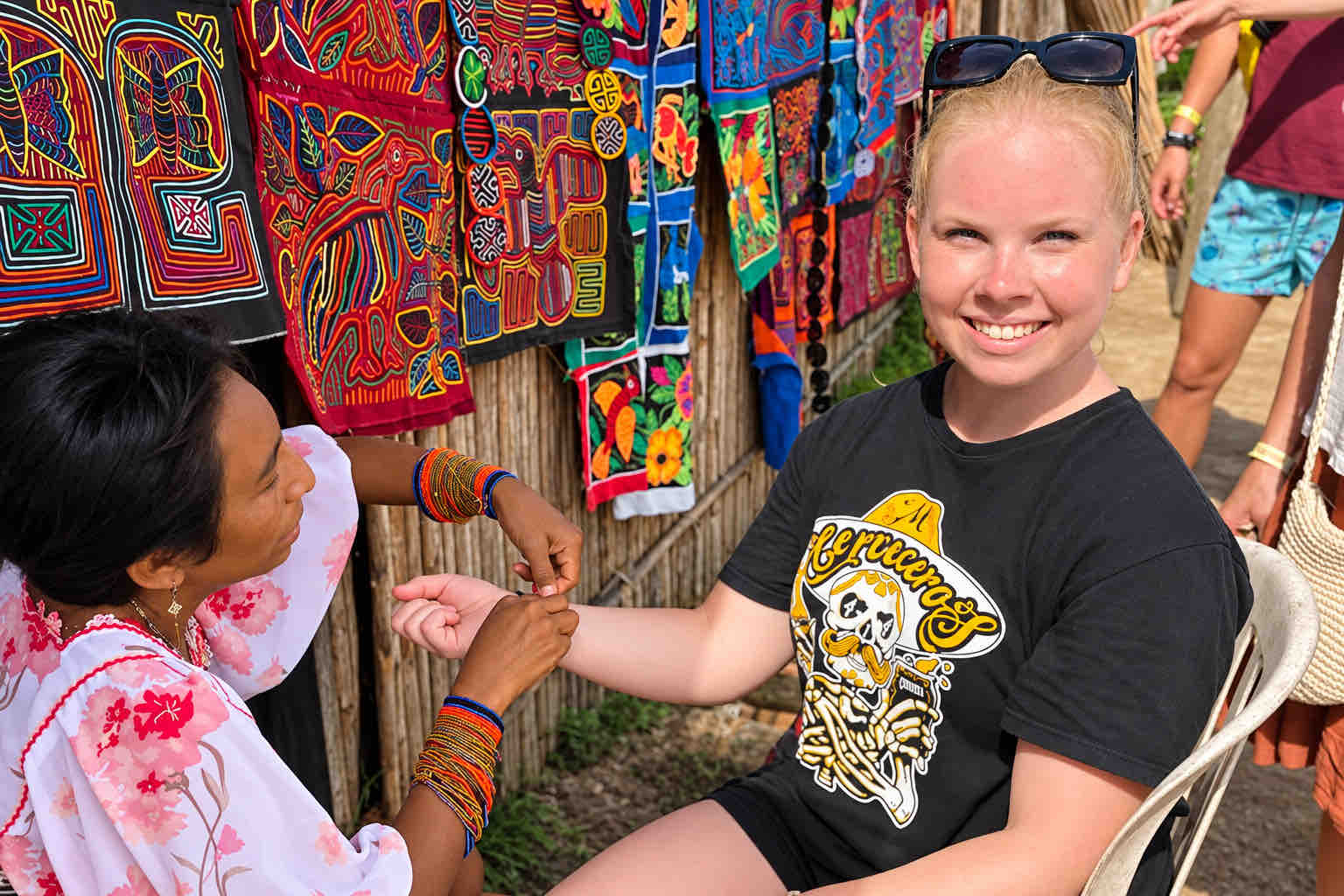 Guna artisan fastening a beaded bracelet on a visitor beside mola textiles