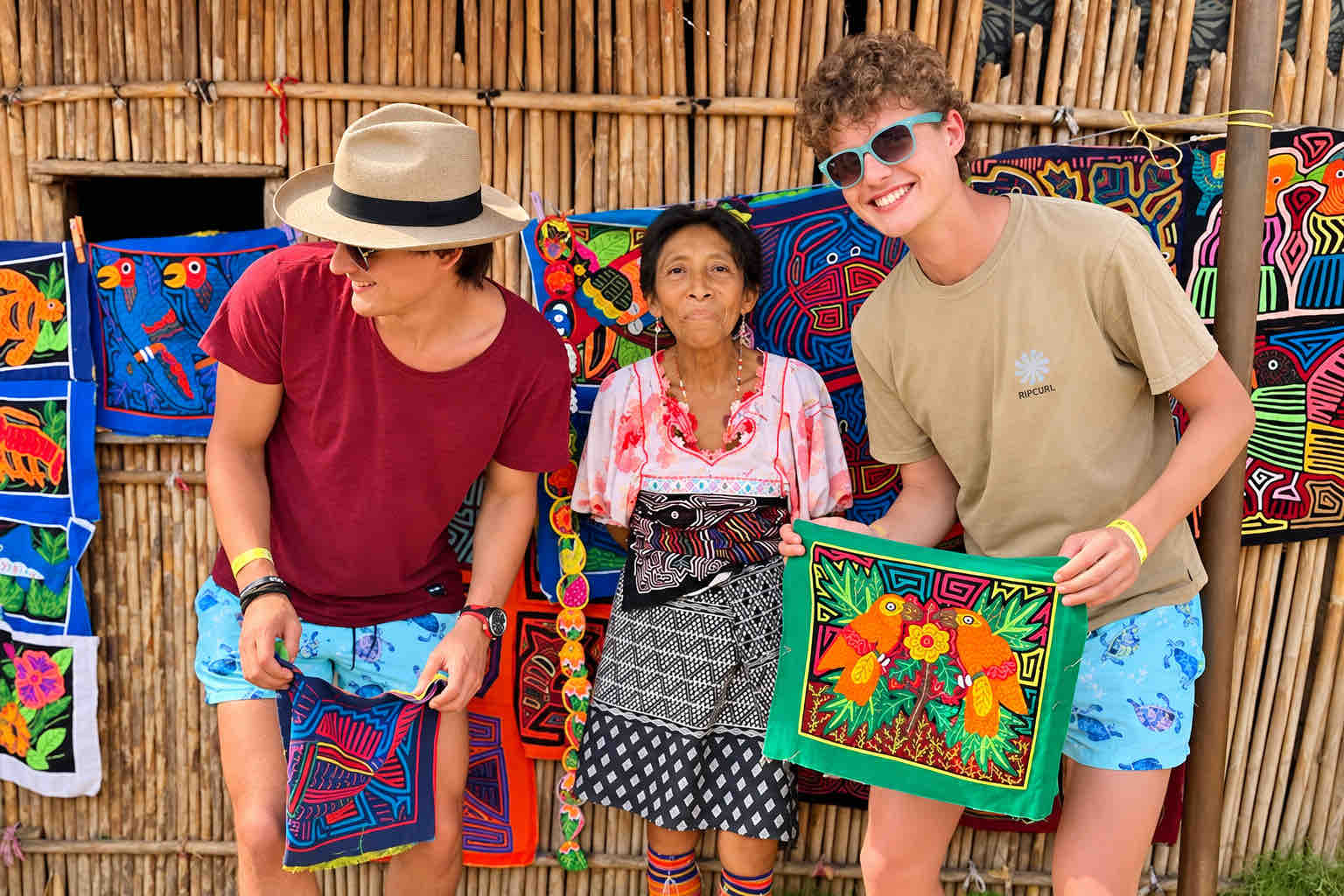 Guna Yala Tour – Mola Textile Visit Two visitors and a Guna host posing in front of colorful mola textiles