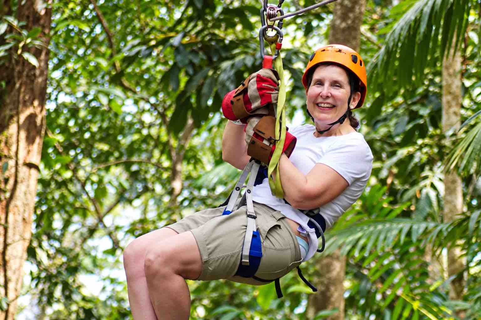 Smiling woman wearing an orange helmet and gloves riding through foliage on the Zipline Panama tour