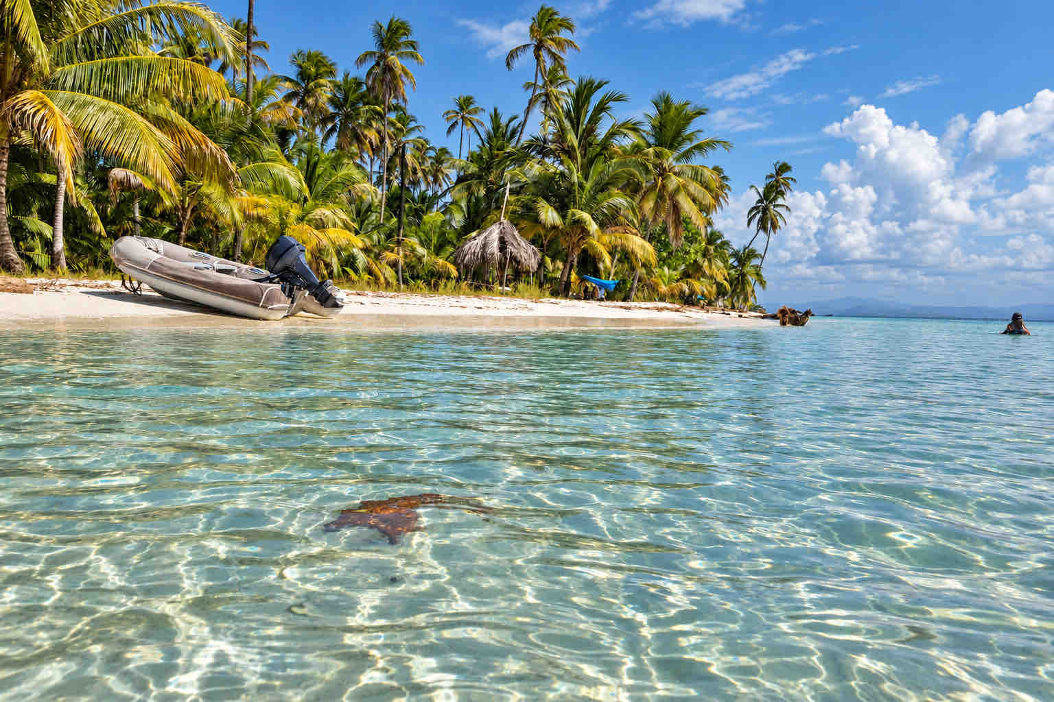 Clear shallow water beside a palm-lined beach and thatched shelter in Cayos Holandeses, San Blas.