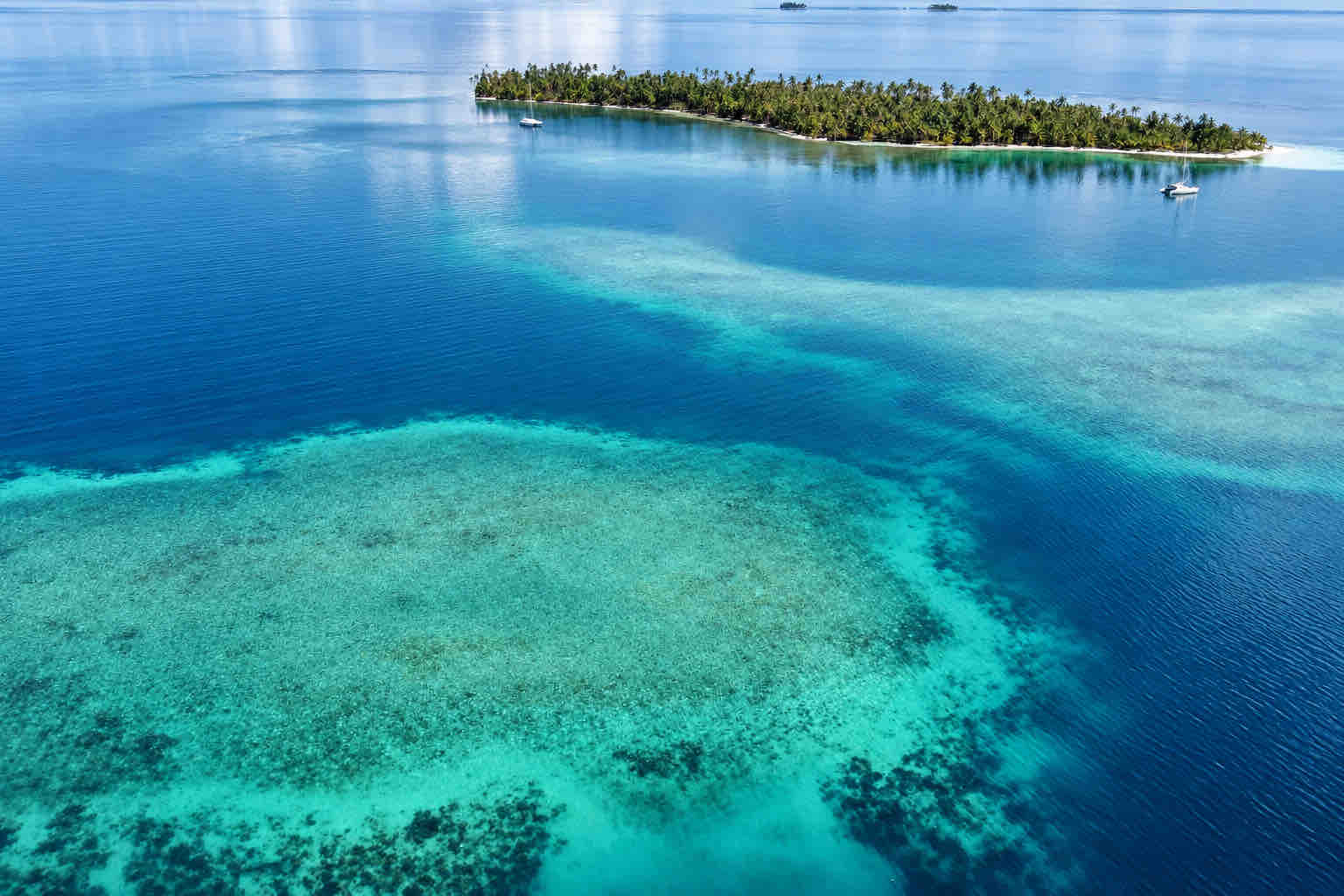 Aerial view of a palm-covered Holandeses island with sailboats and turquoise reef shallows.