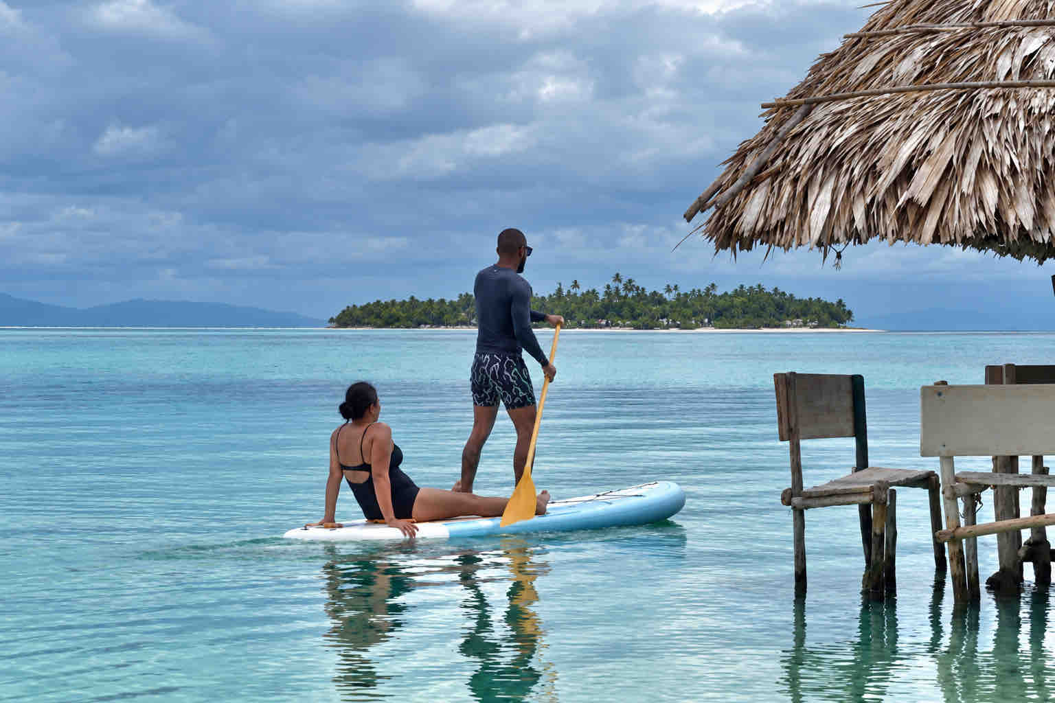 Two travelers on a paddleboard beside a shaded table in the shallow waters of Holandeses.